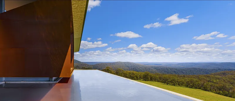 Infinity pool edge with cantilevered corten steel canopy overlooking forested valley under blue sky