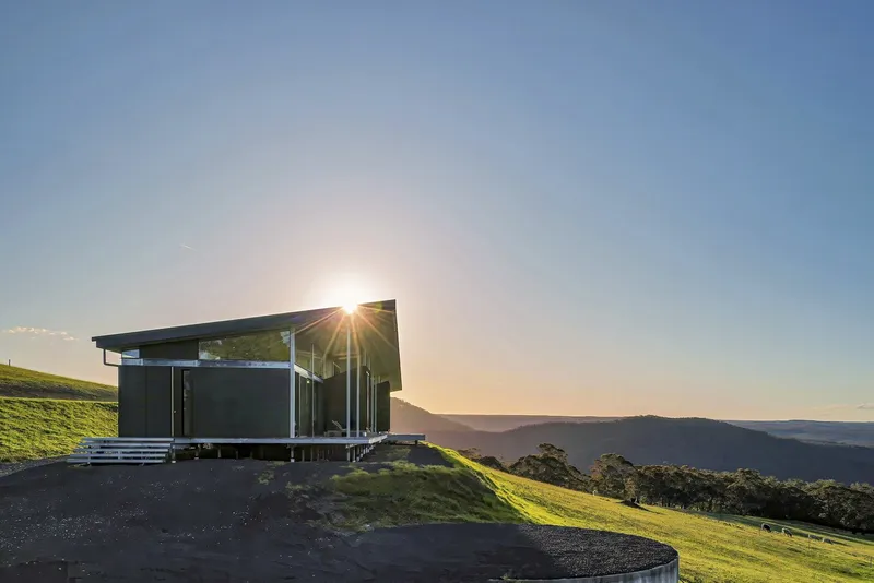Glass-walled pavilion on gravel terrace perched on hillside at sunset