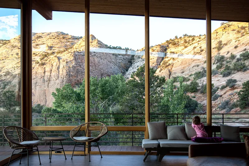 Floor-to-ceiling glazing framing sandstone cliffs and a terrace with seating where a person sits reading