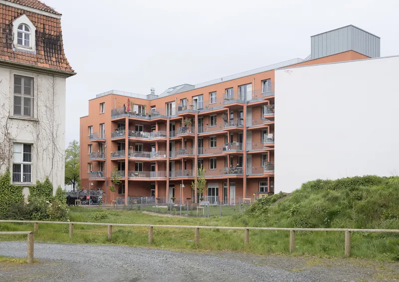 Terracotta brick facade with recessed balconies flanked by older buildings and a grassy field under overcast skies