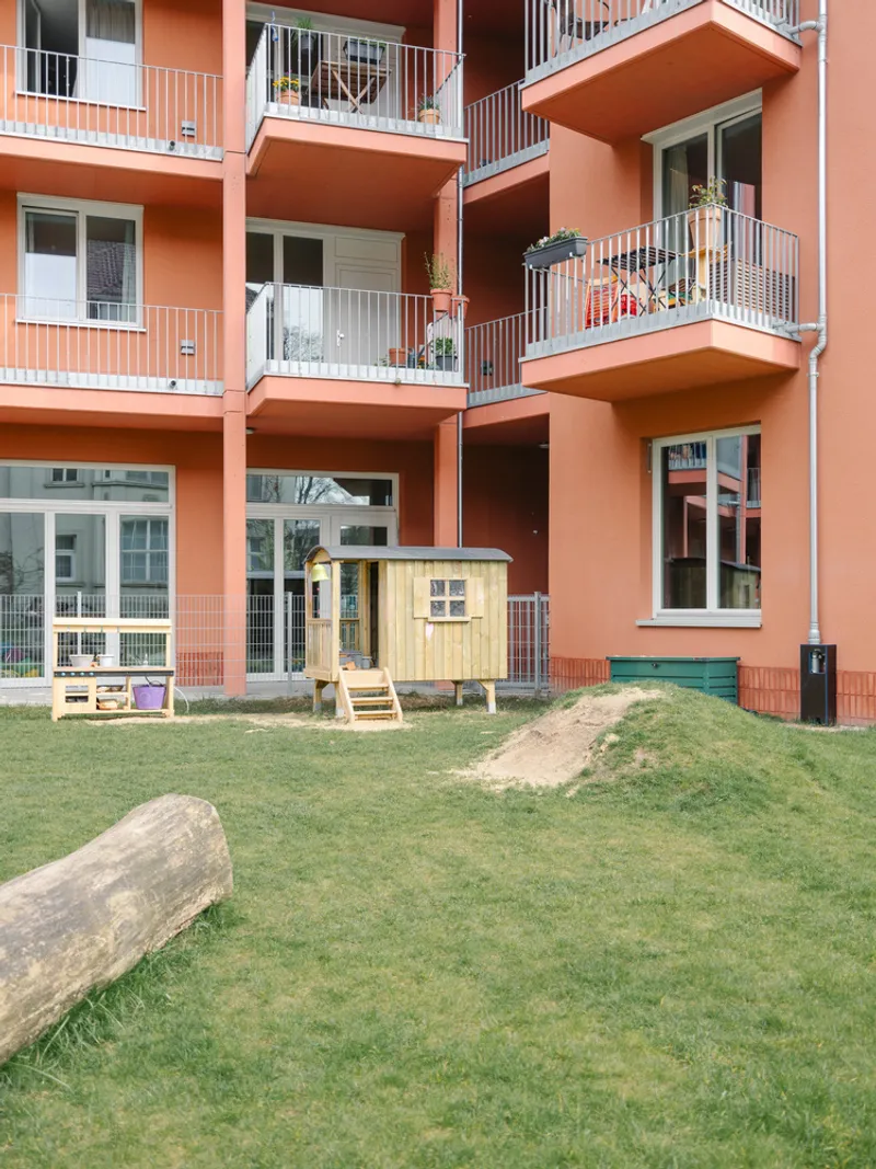 Coral-colored facade with recessed balconies above a shared lawn with a small wooden playhouse