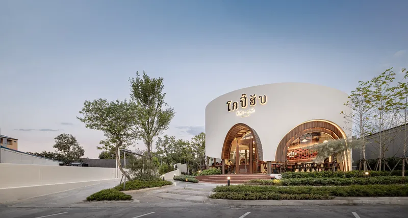 White curved facade with timber-clad arched openings and signage at dusk surrounded by young trees