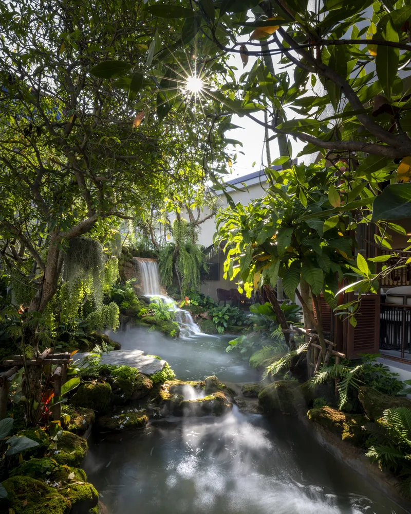 Interior courtyard with waterfall cascading into pond surrounded by ferns and trees with sunlight filtering through canopy