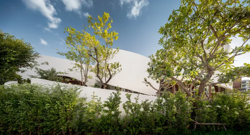 White curved facade walls rising above densely planted gardens with mature trees in daylight