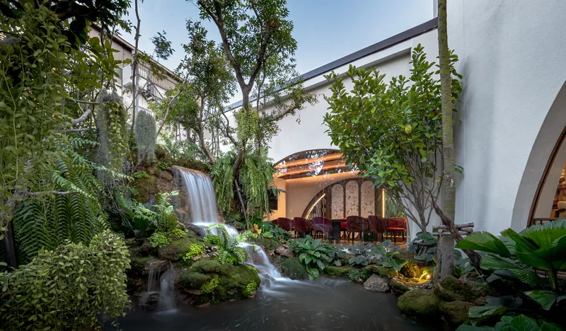 View across pond toward dining pavilion with waterfall and planted landscape at dusk