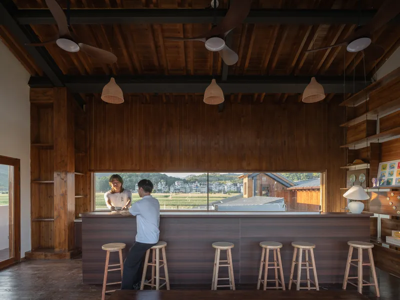 Kitchen counter with timber bar stools and horizontal window framing farmland beyond where two figures stand