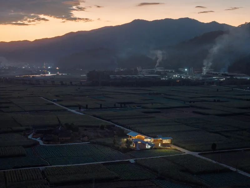 Illuminated residential complex in farmland at dusk with industrial buildings and smoke plumes visible