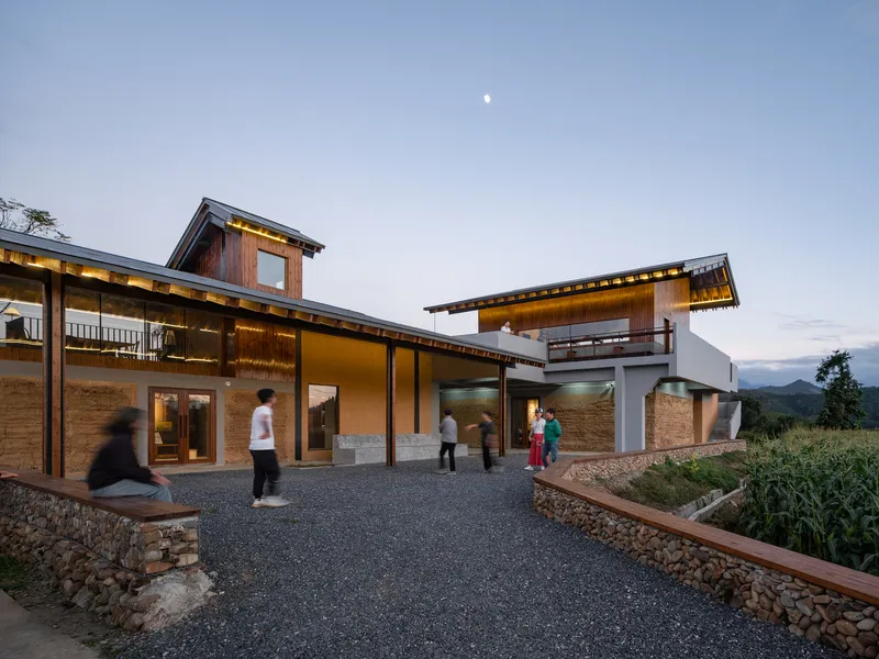 Courtyard entry with gravel surface and people gathering as interior lights glow at twilight