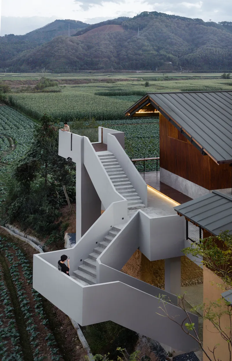 White geometric staircase descending past timber-clad volume toward cultivated fields and distant mountains