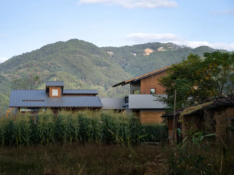 Mixed-material building complex with timber and metal cladding set among cornfields with forested mountains beyond