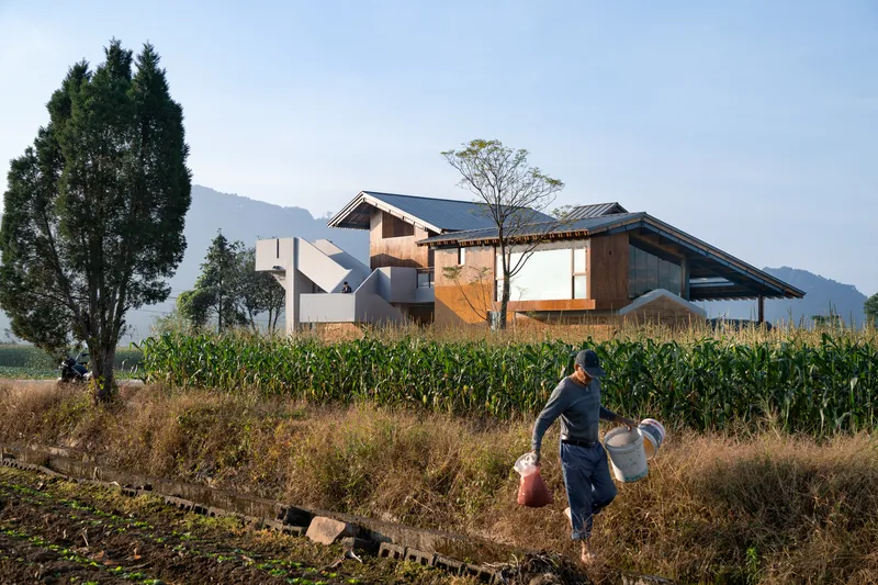 Two figures walking through a cornfield path toward a cluster of timber and metal-roofed volumes