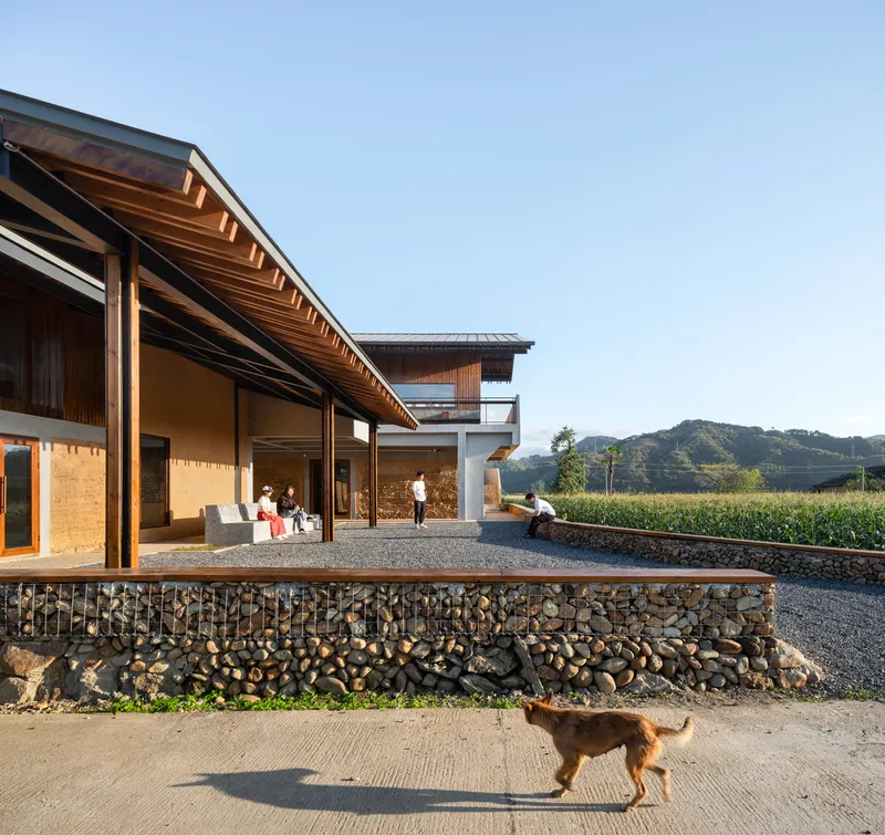 Gabion retaining wall with stone fill supporting a gravel terrace where a dog walks below the eaves
