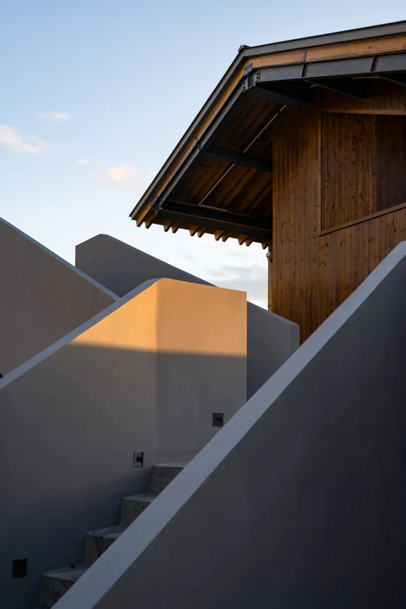Concrete stair ascending through angular rendered walls toward a timber-clad pavilion in evening light