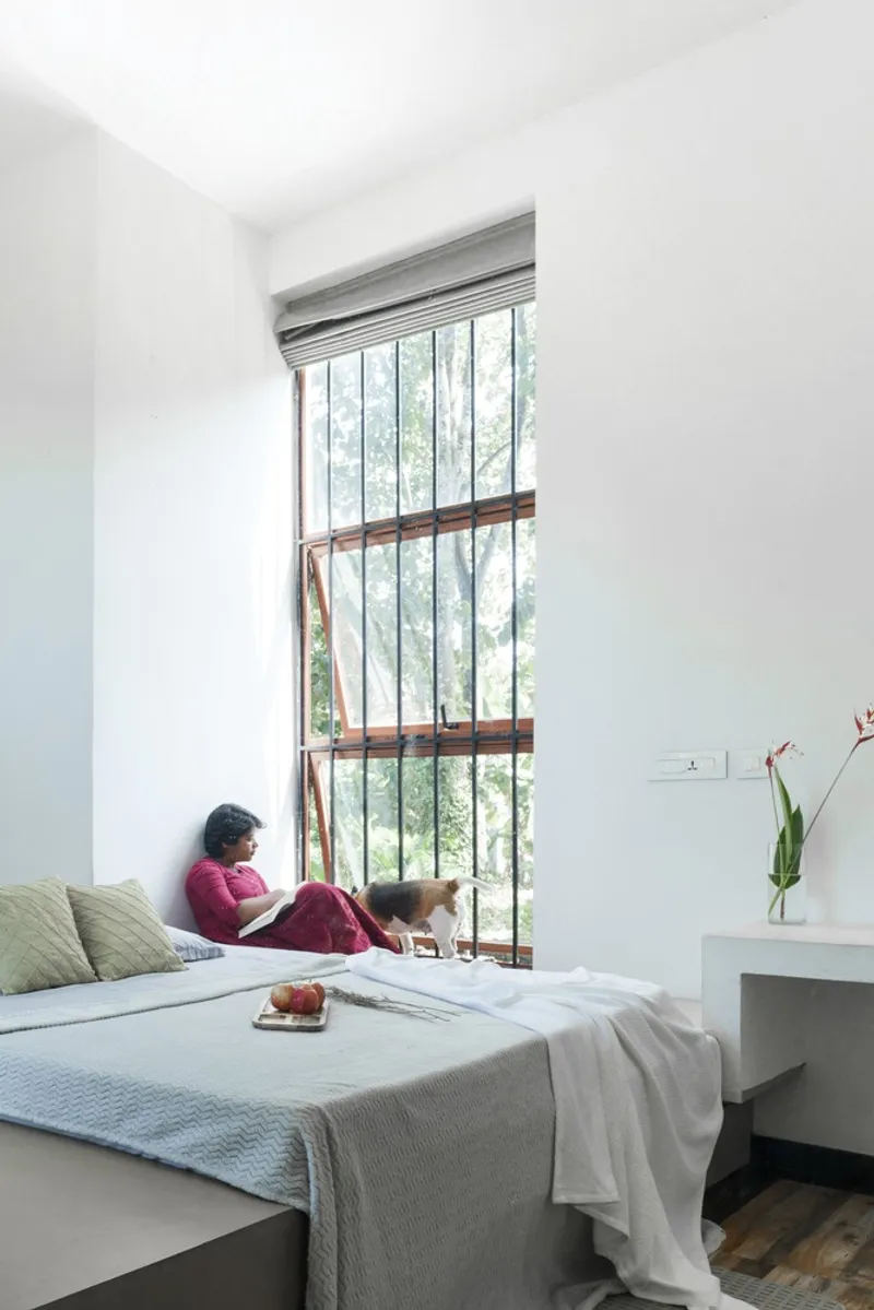 Bedroom with tall steel-framed window overlooking trees and a person reading in the window nook