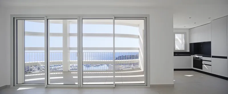 Open-plan living space with kitchen alcove and glazed doors framing harbor view in bright daylight