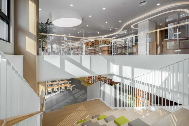 Multi-level interior atrium with white vertical slat railings, library shelving visible above and sunlight streaming through glazing