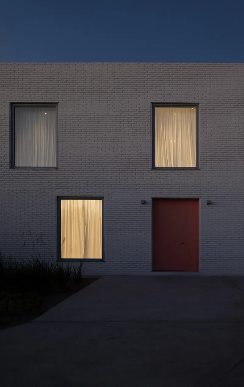 Detail of grey brick facade showing three windows with sheer curtains and a red entry door