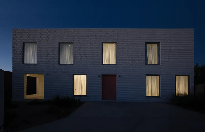 Dark brick facade with illuminated windows and a central red door at dusk