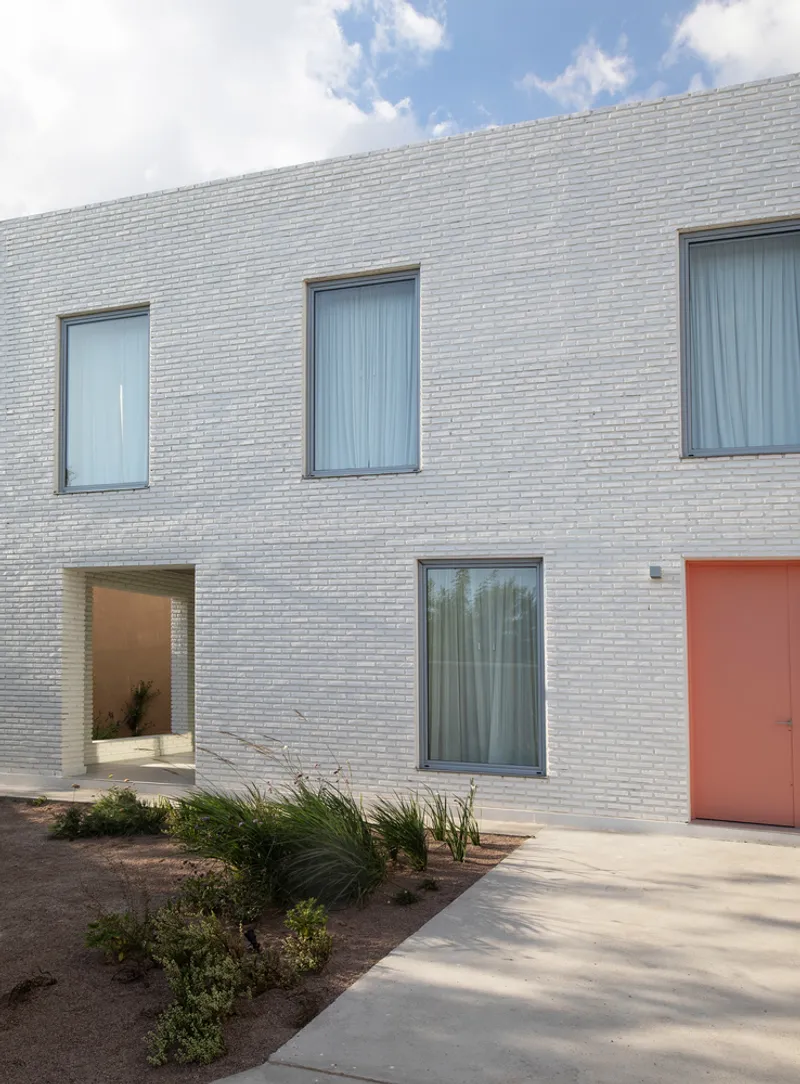 White brick facade with recessed entry and coral door beside planted beds with grasses