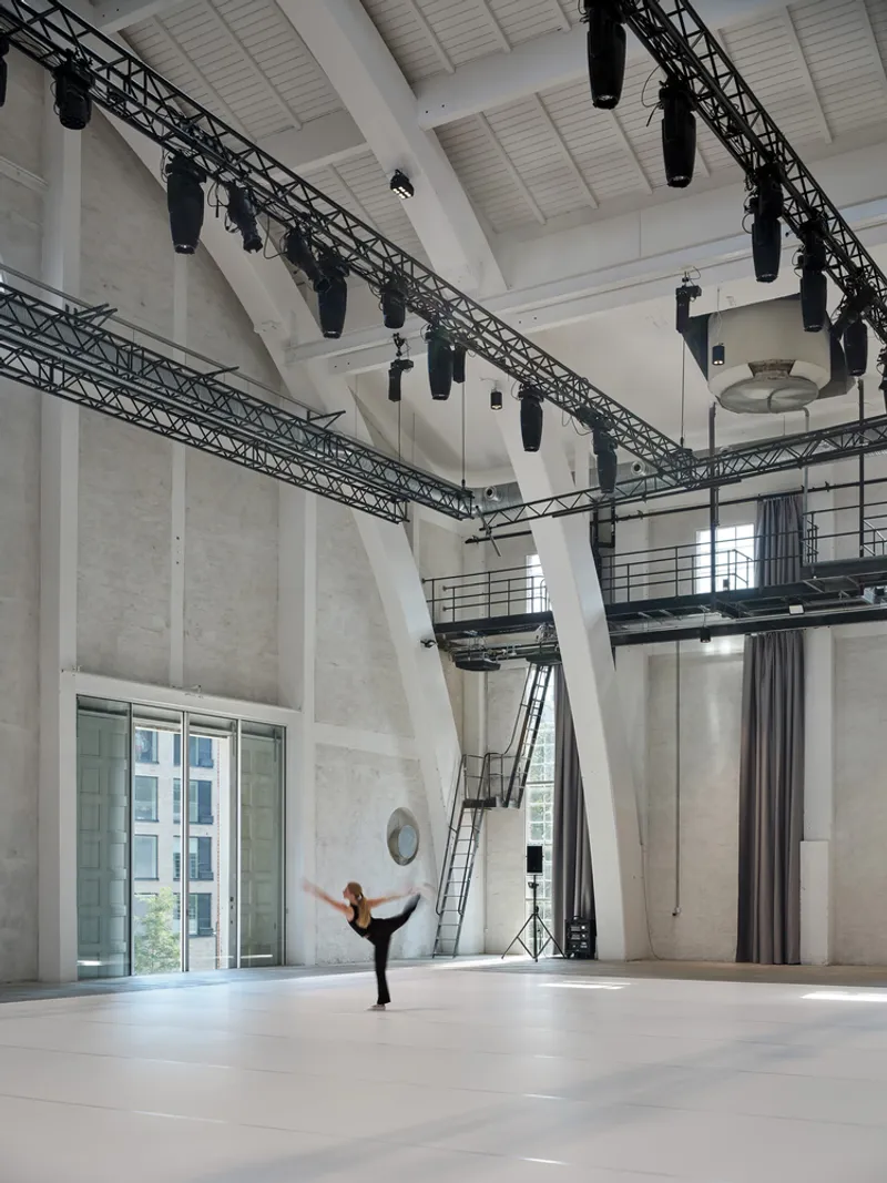 Performance space showing concrete walls, suspended lighting trusses, and a dancer stretching on the floor