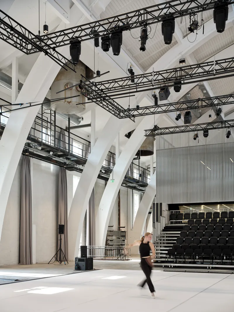 Performance hall interior with white branching columns supporting stage lighting trusses as a figure walks across the floor