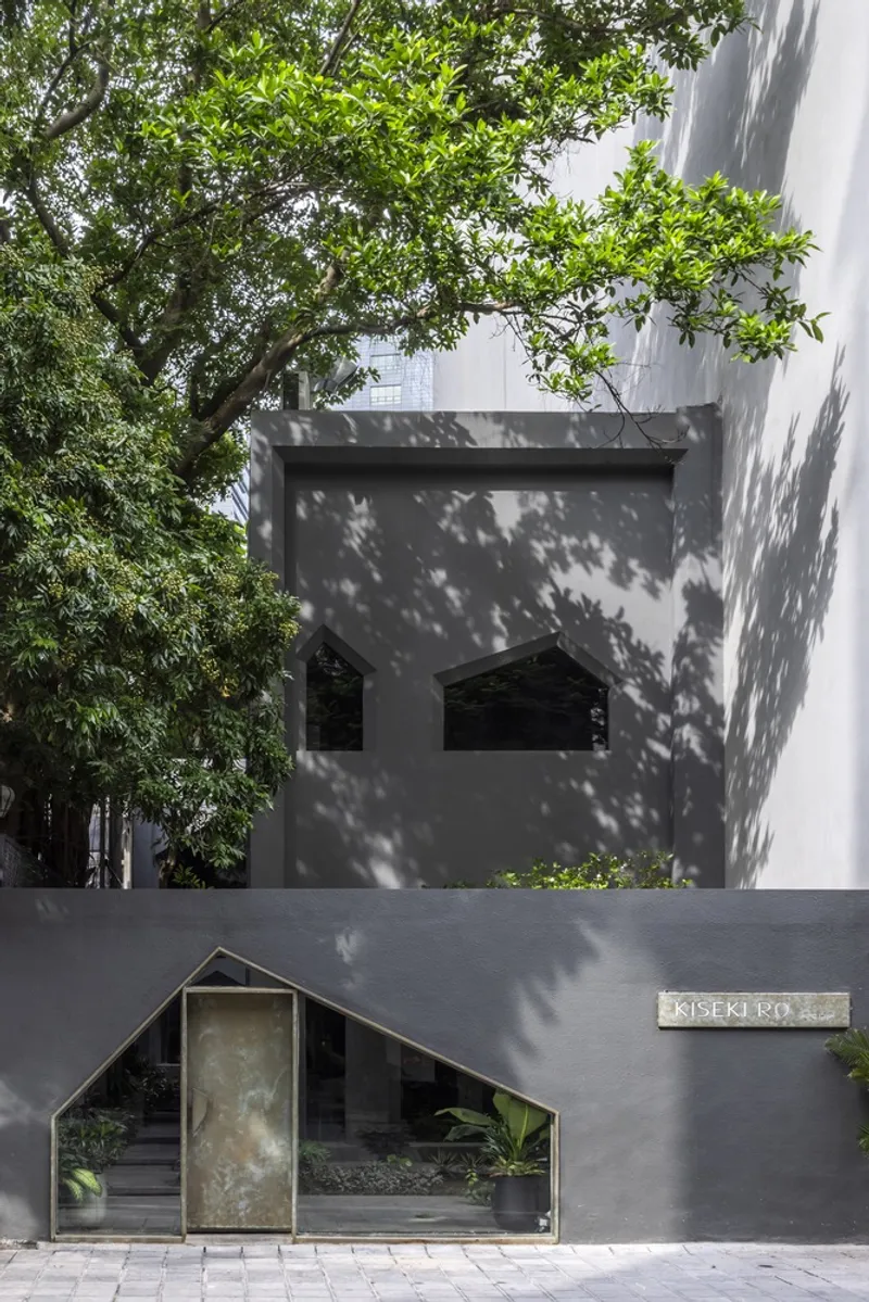 Street entrance with peaked metal gate beneath a cantilevered volume casting tree shadows