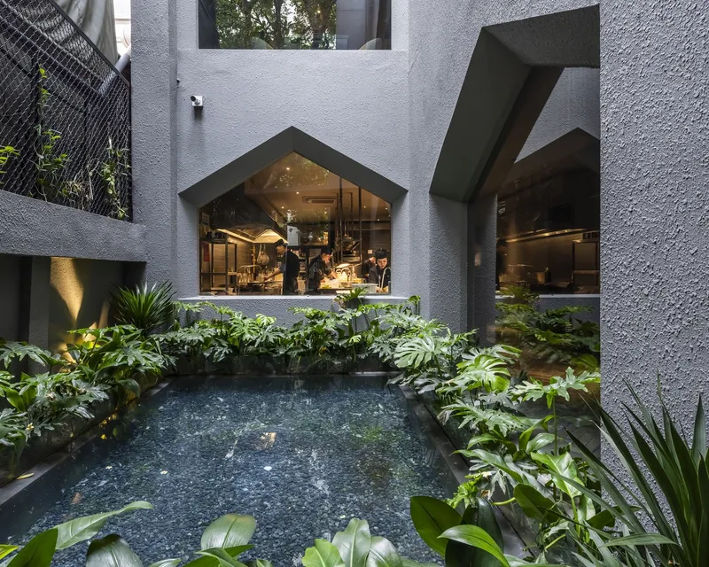Courtyard pool surrounded by planted beds and pentagonal windows looking into lit kitchen space