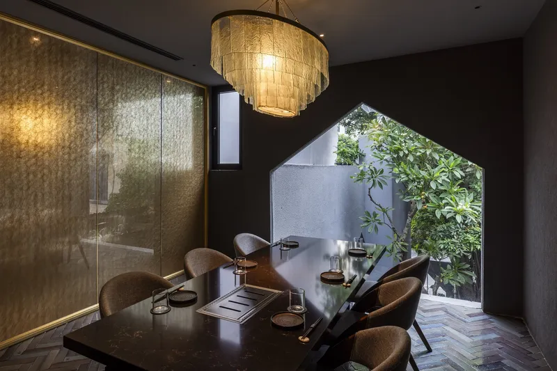 Dining room with tiered pendant light, textured gold wall panels, and pentagonal opening to planted courtyard