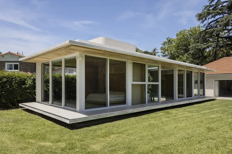 Angled view of the glazed pavilion on a raised plinth surrounded by hedges and lawn