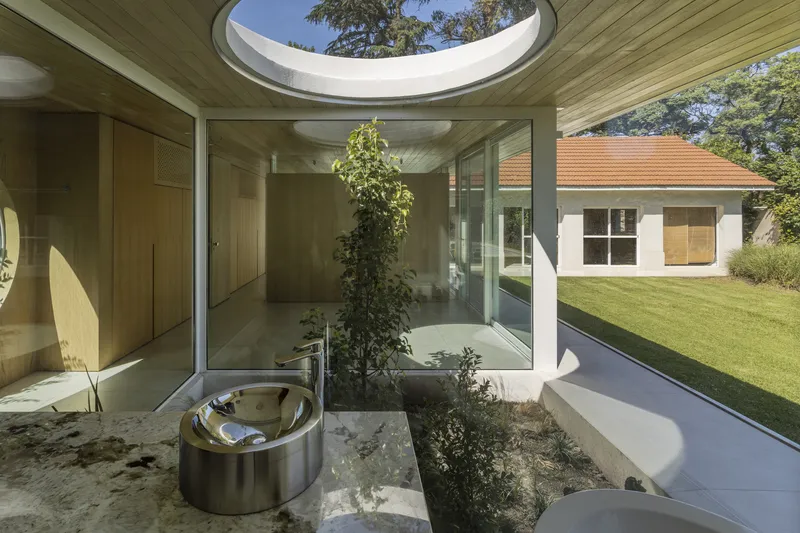Glazed courtyard interior with planted bed, polished metal bathtub, and oval skylight framing trees above