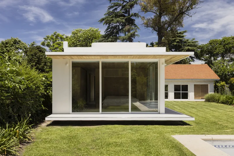 Garden elevation of the pavilion with flat roof and full-height glazing set in a lawn under cloudy sky