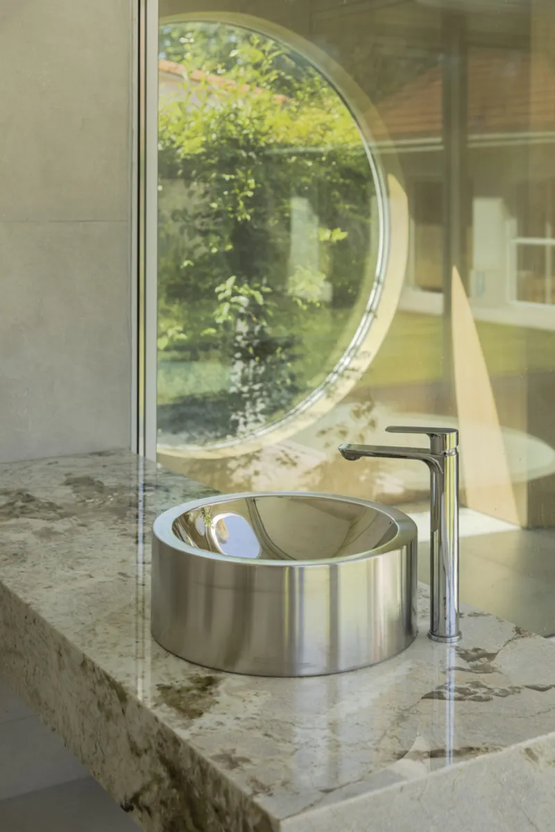 Stainless steel basin on stone countertop reflecting the circular garden window behind glass