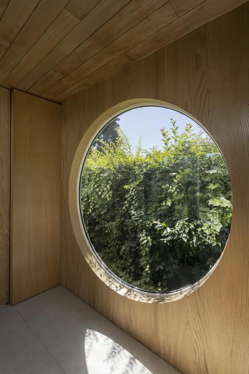 Circular window framed in oak framing views of dense hedge beyond interior timber walls