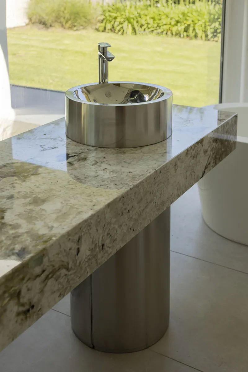 Polished stainless steel vessel sink on a textured stone countertop overlooking a garden