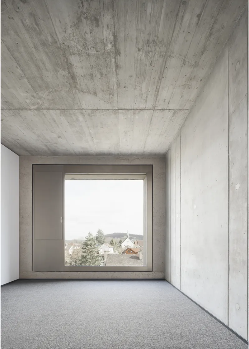 Empty concrete room with square window framing a village view under overcast sky