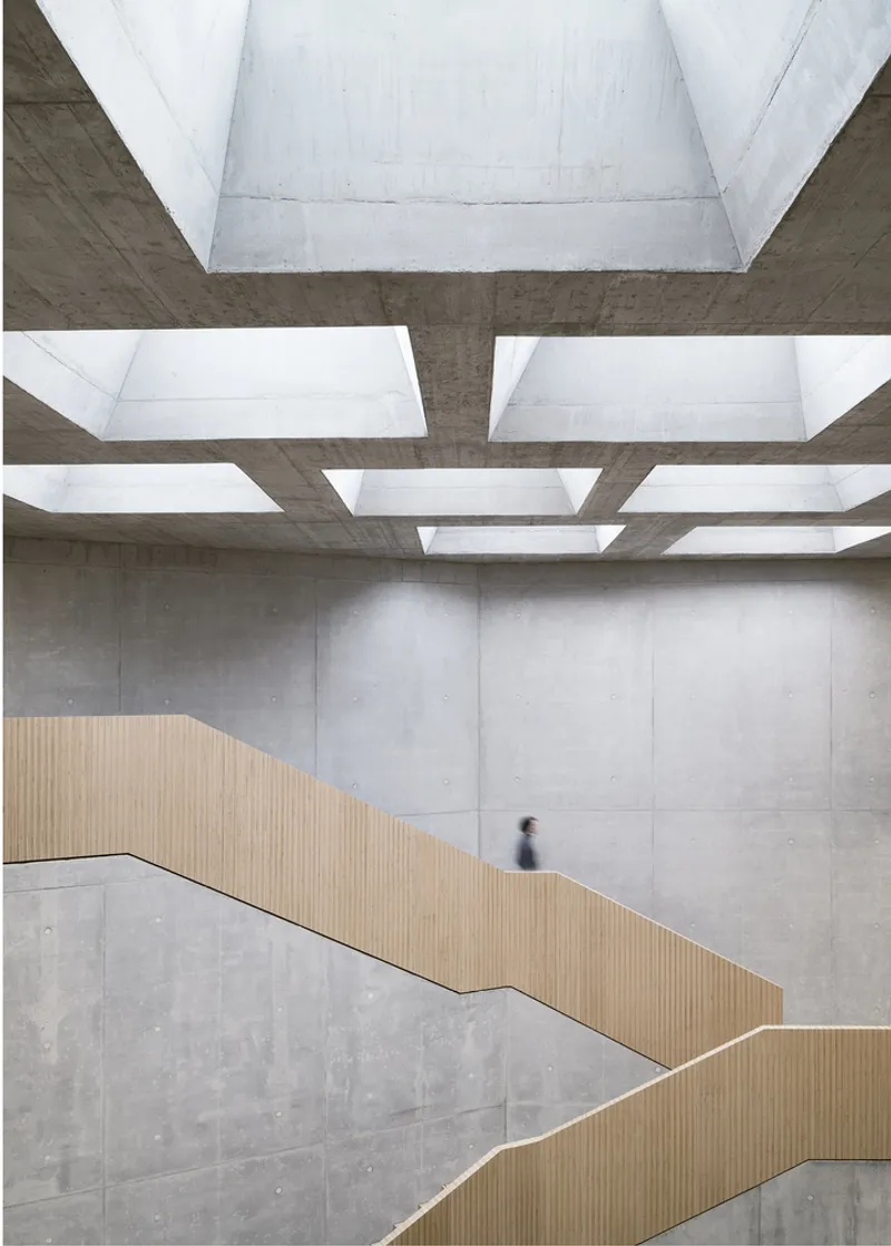 Timber-clad zigzag staircase beneath concrete coffers with rectangular skylights and a person ascending