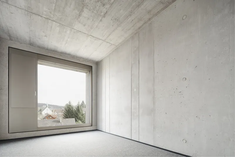 Bare concrete interior with exposed formwork ceiling and square window overlooking rooftops
