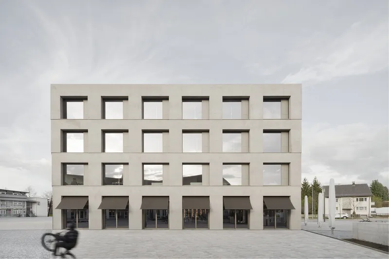Grid facade with deep-set square windows and ground-level awnings as a cyclist passes the plaza
