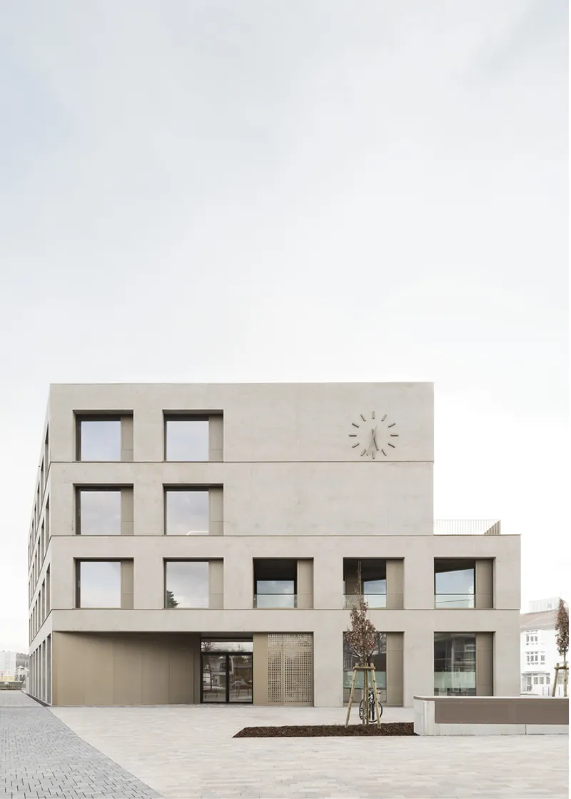 Street view of the beige stone facade with square windows and a wall-mounted clock under overcast sky