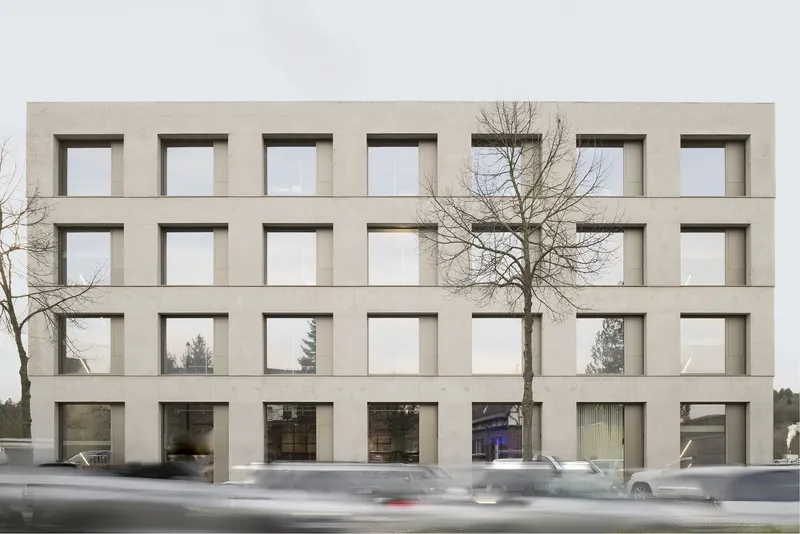 Facade with recessed square windows partially obscured by a bare winter tree and passing traffic
