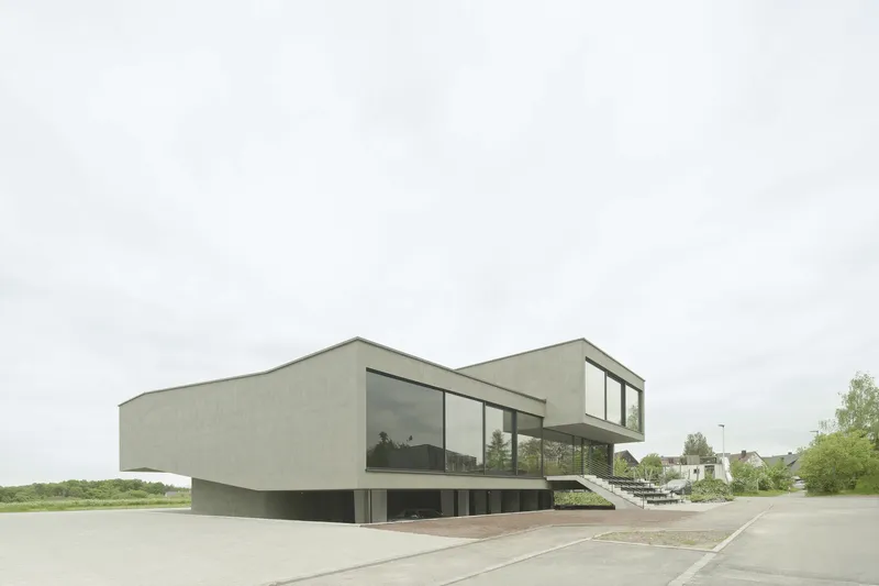 Elevated concrete structure with glazed openings and recessed ground floor under cloudy sky