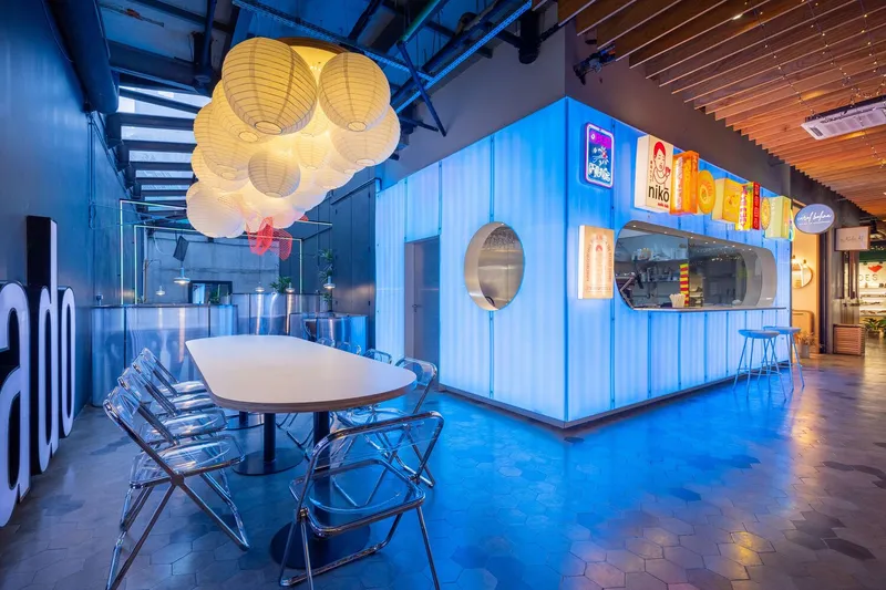 Food hall interior with corrugated metal kiosk, cluster of paper lanterns, and blue ambient floor lighting