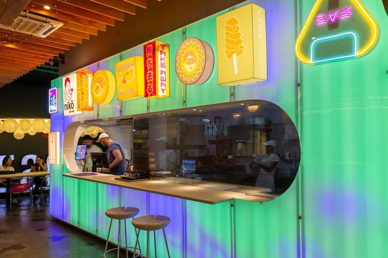 Service counter with oval glass opening, backlit signage above, and wooden bar stools along corrugated metal base