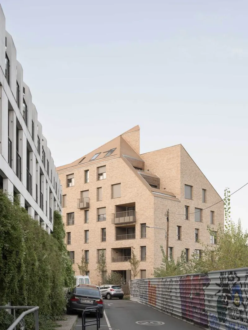 Gabled brick volume viewed from the street with young trees and a neighboring white facade