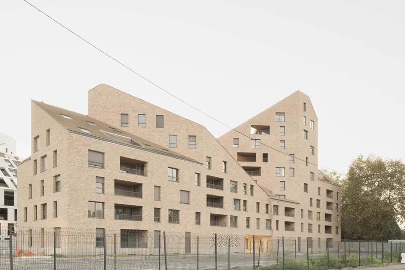 Multi-unit residential building with pale brick facade and angled rooflines behind a perimeter fence