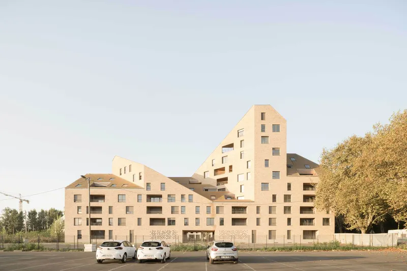 Stepped massing of pale brick volumes rising against a clear evening sky with parked cars below