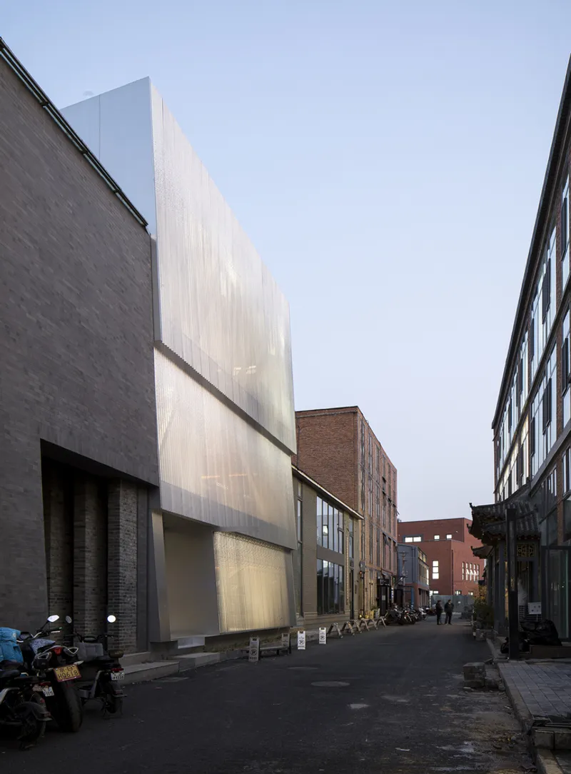 Illuminated translucent facade along narrow street with pedestrians and parked motorcycles at dusk