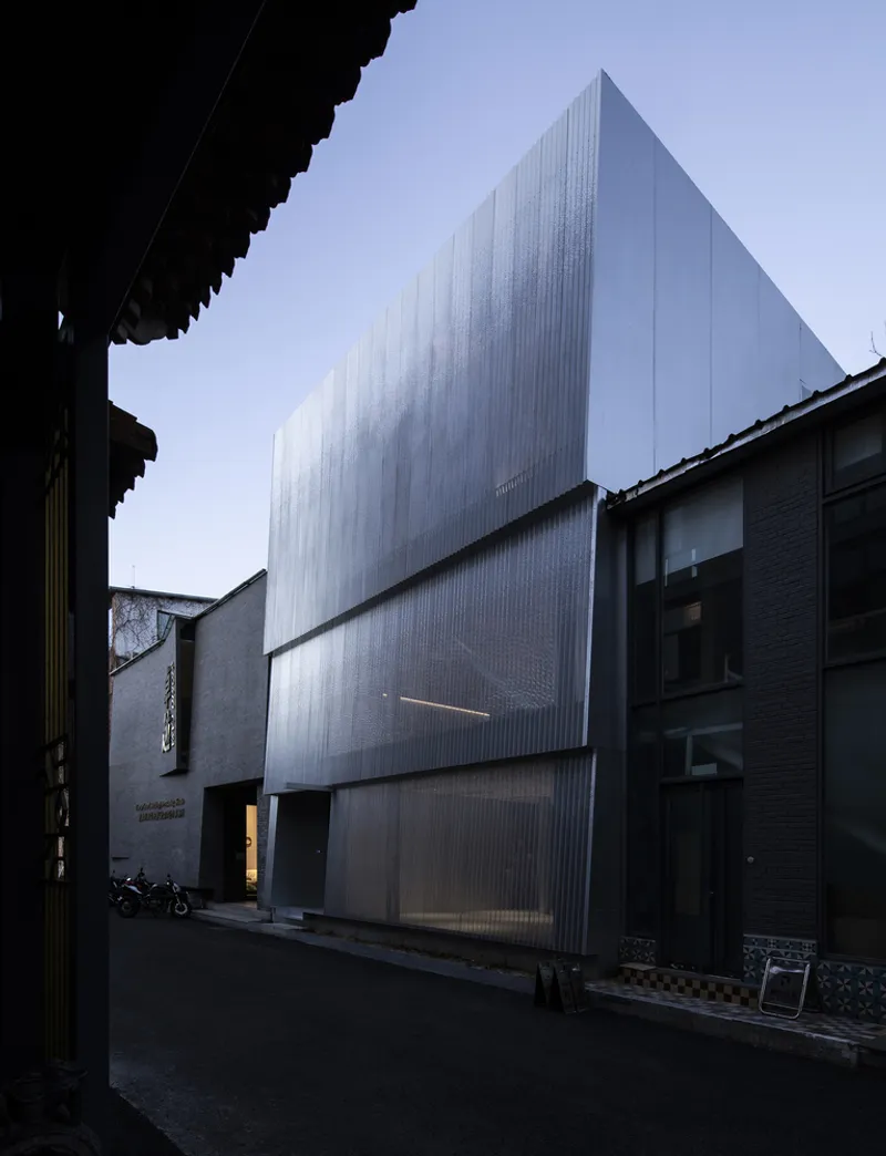 Corrugated metal facade rising above low neighboring roofs in a narrow lane at dusk