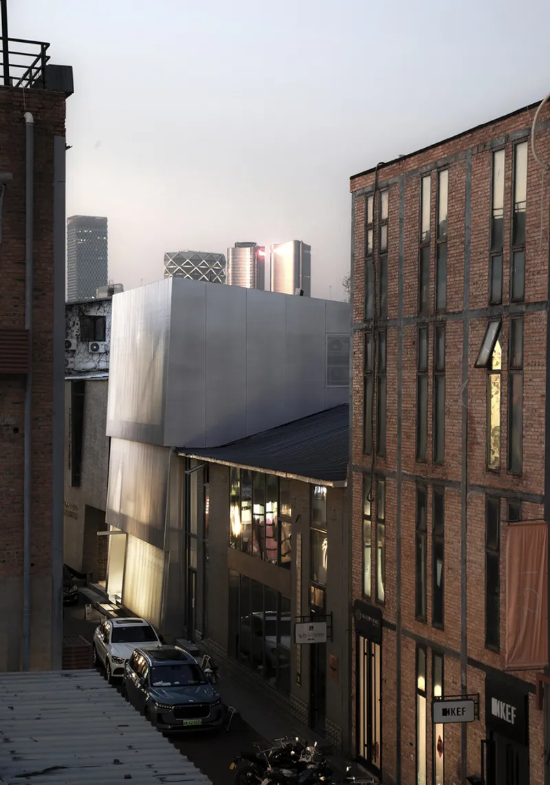 Narrow alley view showing the curved metal facade wedged between brick buildings at dusk with lit interiors