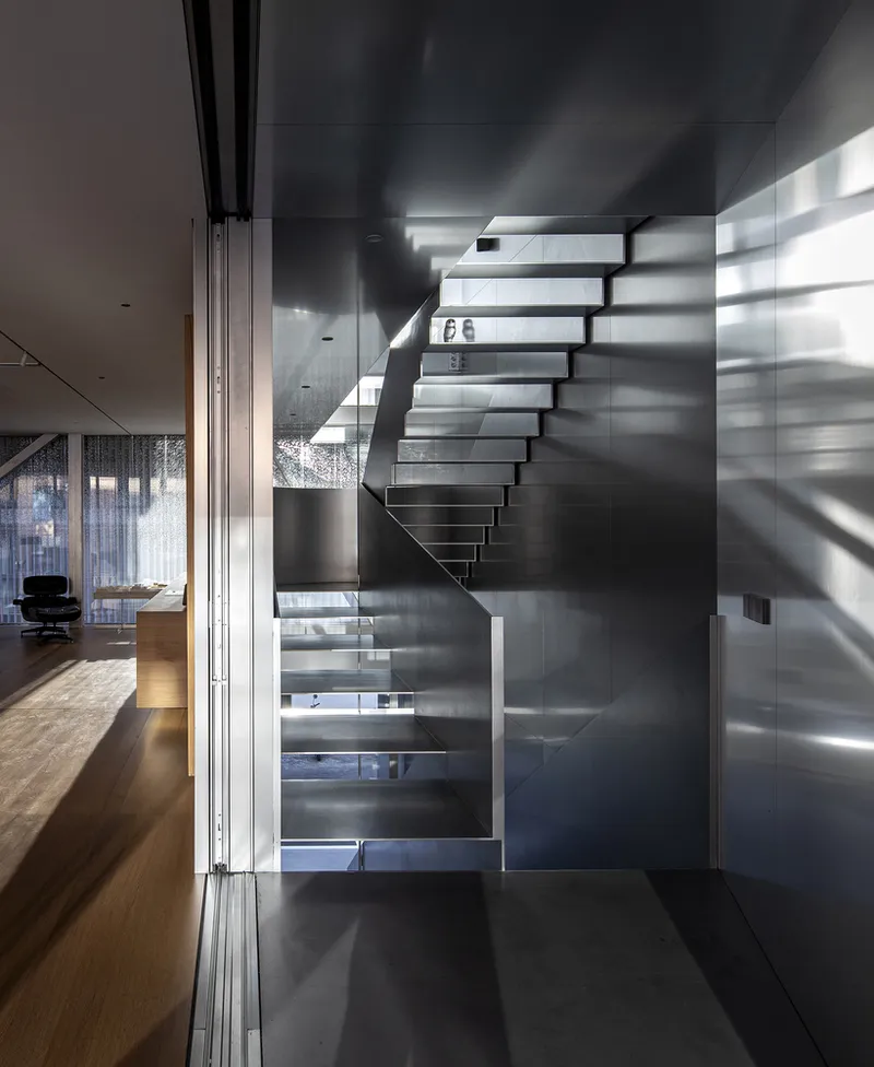 Cantilevered steel staircase with open treads and glass railing casting striped shadows across grey walls